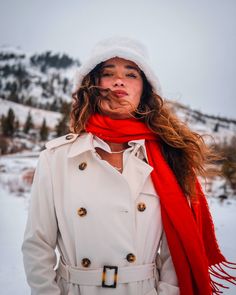 a woman wearing a white trench coat and red scarf standing in the snow with her eyes closed