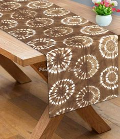 a wooden table with a brown and white table cloth on it's edge, in front of a flower vase