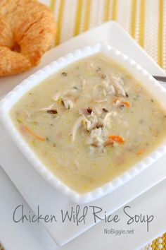 a white plate topped with a bowl of chicken wild rice soup next to a croissant