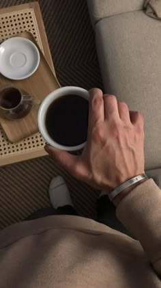 a man holding a cup of coffee on top of a couch next to a tray