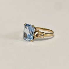 Beautiful 1940s era vintage 10k rosy yellow gold ring featuring a swiss blue topaz stone. The radiant blue topaz stone is radiant cut and secured by four prongs. The ring's shoulders are elegantly cinched with milgrain and repousse design details. Lovely piece of fine vintage jewelry, circa 1940! ERA - Circa 1940s / Retro METAL / MATERIAL - 10k gold, blue topaz MARKINGS / HISTORY - 10k SIZE / MEASUREMENTS - Size: 6 , Swiss Blue Topaz stone: approximately 10 x 8 mm, Weight: 2.15 grams, Shank: Tap Classic Light Blue Topaz Ring For Formal Occasions, Vintage Yellow Gold Rings With Blue Topaz, Vintage Yellow Gold Blue Topaz Rings, Classic Light Blue Topaz Ring For Anniversary, Classic Topaz Birthstone Ring, Classic Blue Topaz Ring, Vintage Blue Topaz Ring In 14k Gold, Vintage Solitaire Topaz Ring, Vintage Blue Sapphire Ring Stamped 14k