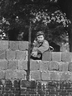 black and white photograph of a man leaning on a brick wall with trees in the background