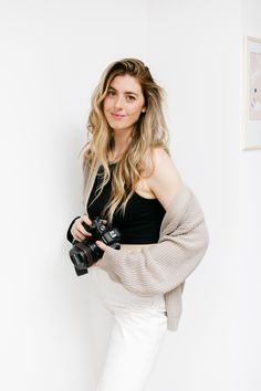 a woman is holding a camera and posing for a photo in front of a white wall