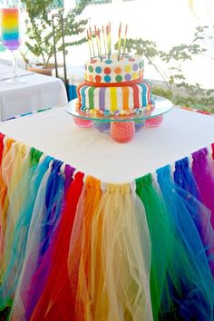 there is a cake on the table with colorful tulle skirt around it and balloons in the background