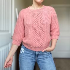 a woman wearing a pink sweater and jeans standing in front of a door with her hands on her hips