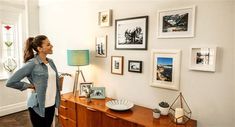 a woman standing in front of a dresser with pictures on the wall behind her and a lamp next to it