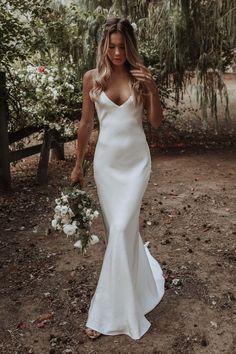 a woman in a white wedding dress is walking through the woods with her hand on her hip