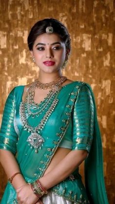 a woman in a green and white sari with jewelry on her neck, standing against a gold wall