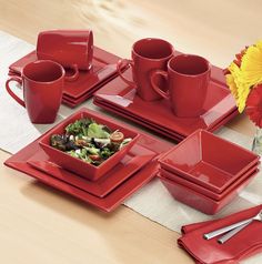 a table set with red dishes, cups and utensils in front of flowers