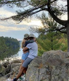 a man and woman hugging on top of a rock near the water with trees in the background