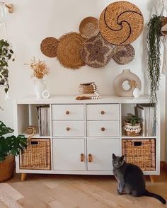 a black cat sitting on top of a wooden floor next to a white cabinet with wicker baskets