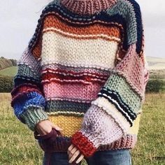 a woman standing in a field wearing a multicolored sweater