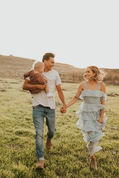 a man and woman holding hands while walking through the grass with a baby in their arms