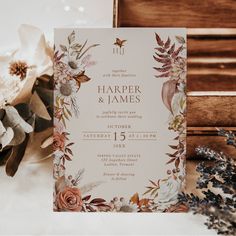 a wedding card with flowers and greenery on it next to a flower arrangement in front of a wooden box