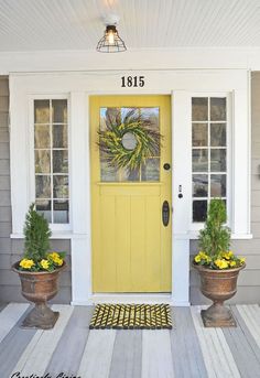 the front door is painted yellow and has two potted plants next to it on the porch