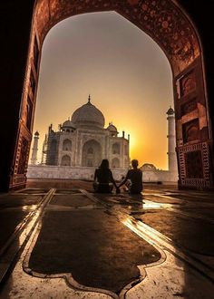 two people sitting on the ground in front of an arch with a sunset behind them