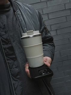 a man holding a coffee cup and cell phone in his hand while standing next to a brick wall