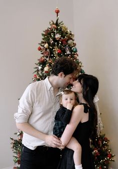 a man and woman kissing while holding a small child in front of a christmas tree