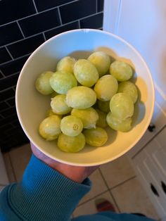 a person holding a white bowl filled with green fruit on top of a tiled floor