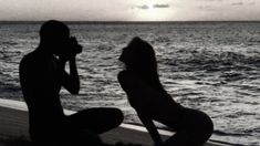 a man and woman sitting on the beach next to each other, with water in the background