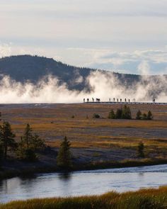 steam rises from the ground as people walk across an open field with mountains in the background