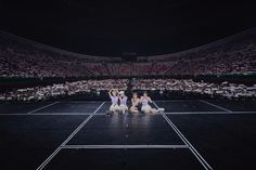 three women sitting on the ground in front of a crowd at a tennis match, all dressed in white