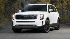 the front end of a white suv driving down a road near some trees and bushes