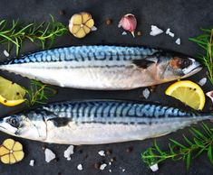 two fish with lemon slices and herbs on a black surface next to some seaweed