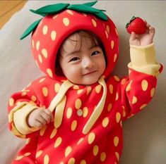 a baby in a strawberry costume is holding up a strawberries on his hand and smiling at the camera