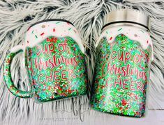 two christmas mugs sitting next to each other on a furry surface with the words cup of christmas cheer printed on them