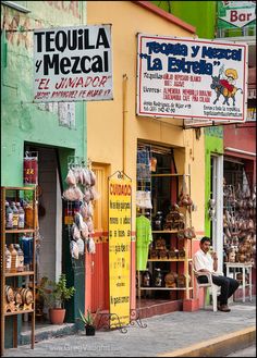 a man sitting on a bench in front of a store with many items hanging from it's windows