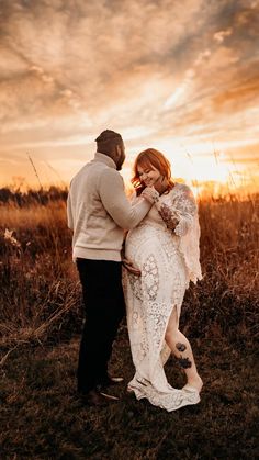 a man and woman standing next to each other in a field with the sun setting behind them