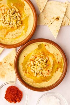 two bowls filled with hummus next to tortillas and pita chips on a white surface