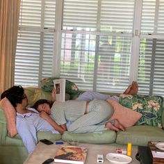 two people laying on a couch in front of a window with the blinds pulled down