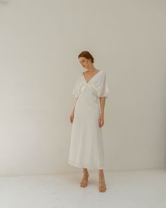 a woman standing in front of a white wall wearing a white dress and tan sandals