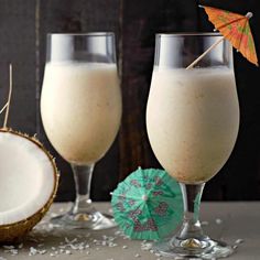 two glasses filled with white liquid next to a coconut and an umbrella on a table