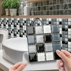 two hands are holding up a mosaic tile design in front of a white sink and toilet