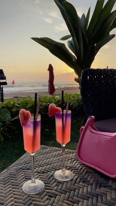 two glasses with drinks sitting on a table near the ocean at sunset or sunrise,