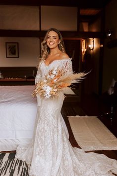 a woman standing in front of a bed wearing a wedding dress with off the shoulder sleeves