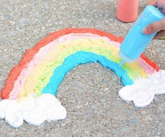 someone is painting a rainbow with white and blue icing on the ground next to a cup