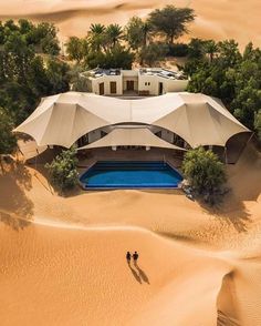 an aerial view of two people standing in the sand near a large tent and pool