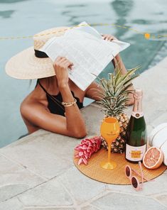 a woman sitting next to a pool with a pineapple, wine and glasses on it