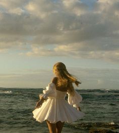 a woman in a white dress standing on the beach looking out at the ocean with her back to the camera