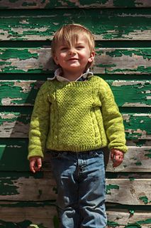 a little boy standing in front of a wooden wall wearing a green sweater and blue jeans