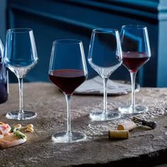 four wine glasses are lined up on a table with some food in front of them