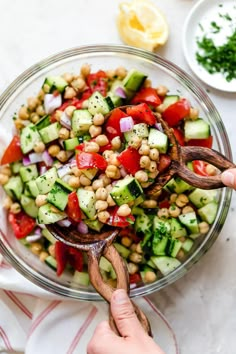 a person holding a wooden spoon over a bowl filled with cucumber and chickpeas