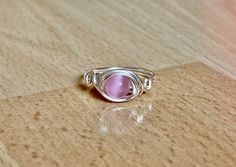 Gemstone: Cats Eye. Gemstone Color: Pink. Band Color: Silver Band Wire: Silver plated copper. Enamel Coating to prevent tarnish and color change. This fun ring has a 6mm glowing pink cat's eye wrapped in 20 gauge silver plated wire. The wire is silver plated with a tarnish resistant coating to prevent wear and color change. This ring is simple and dainty with a swirl design on either side of the stone. Cat's eye is a beautiful, man made stone with a brilliant flash that dances in the light. Chec Handmade Pink Toe Ring Jewelry, Handmade Pink Toe Ring, Nickel Free Pink Promise Ring, Adjustable Pink Moonstone Ring, Adjustable Pink Moonstone Round Ring, Adjustable Pink Round Moonstone Ring, Adjustable Pink Moonstone Ring As Gift, Adjustable Pink Crystal Birthstone Ring, Eye Ring Silver