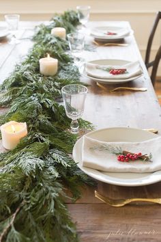 the table is set for christmas dinner with candles and greenery on it, along with white plates