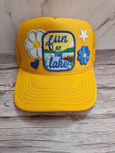 Lake Day Patch Trucker Hat The front of this hat is embellished with patches including one that says fun at the lake, flowers and a heart. This hat is perfect for a day out at the lake or day on the boat. The blue and yellow colors are perfect for summer! The hat has a foam front panel and a mesh back with and adjustable strap in the back. Fun Spring Hats For Outdoor Activities, Fun Spring Outdoor Hats, Fun Outdoor Hat For Spring, Playful Yellow Hats For Spring, Playful Yellow Hat For Spring, Fun Outdoor Spring Hat, Playful Yellow Spring Hat, Retro Summer Hats For Outdoor Activities, Yellow Trucker Hat Baseball Cap For Spring