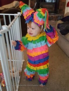 a toddler in a colorful costume standing next to a white gate and looking at the camera